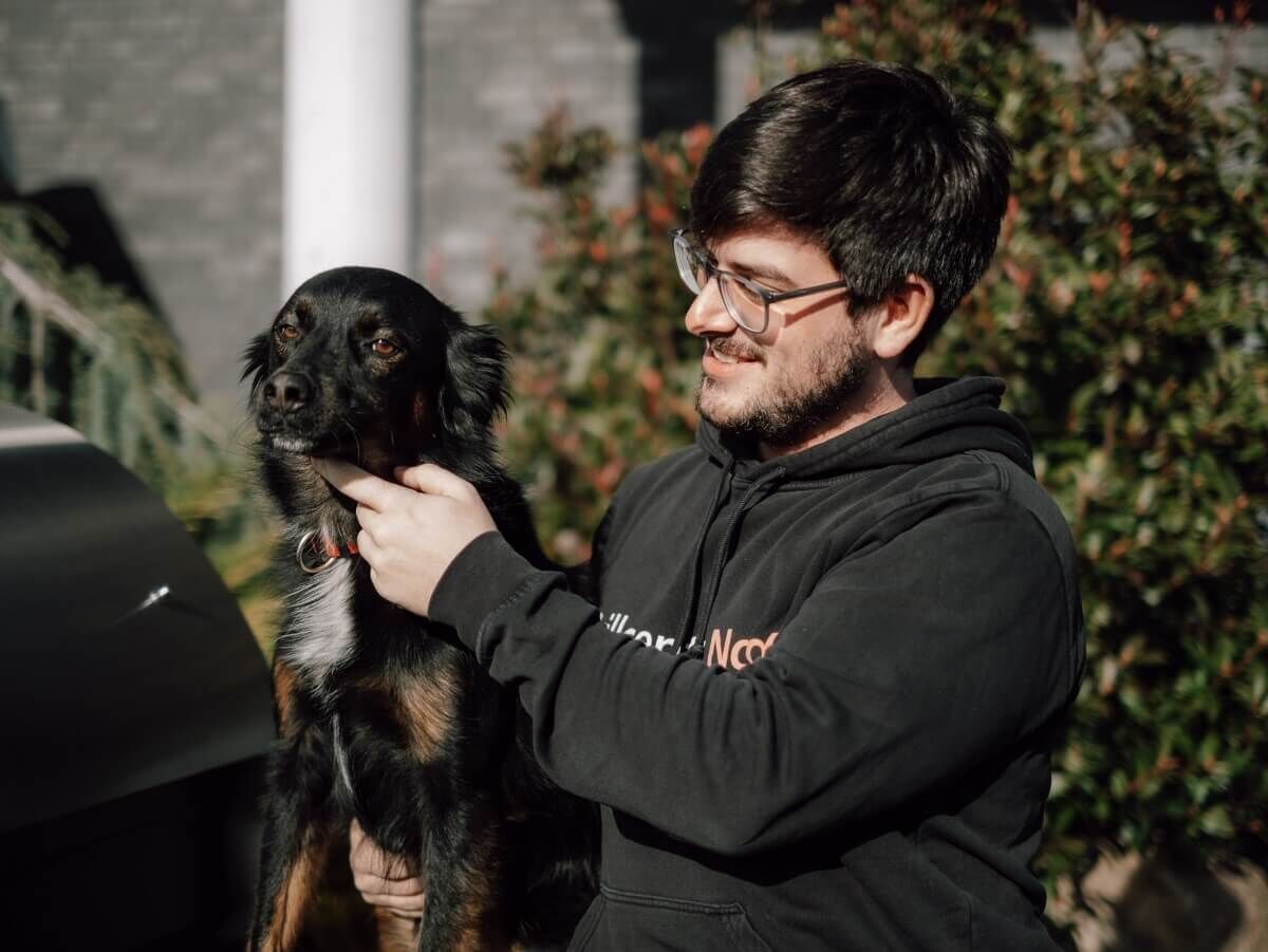 mann-mit-schwarzem-hund-im-arm-trägt-schwarzen-hoodie-und-brille-steht-im-sonnenschein-vor-grünem-hintergrund