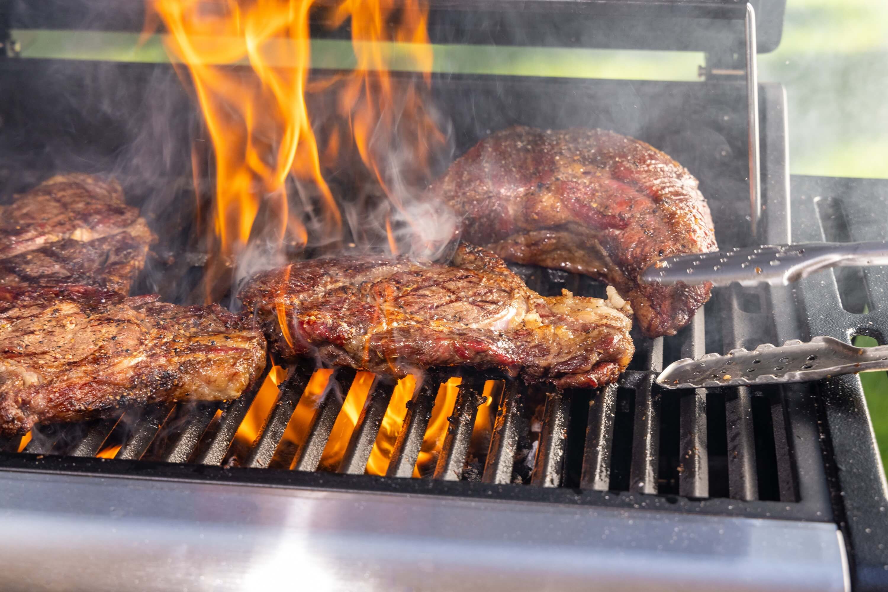 steaks-auf-dem-gasgrill-mit-offenen-flammen-und-rauch-zange-greift-nach-saftigem-fleisch-grillrost-aus-edelstahl-in-nahaufnahme