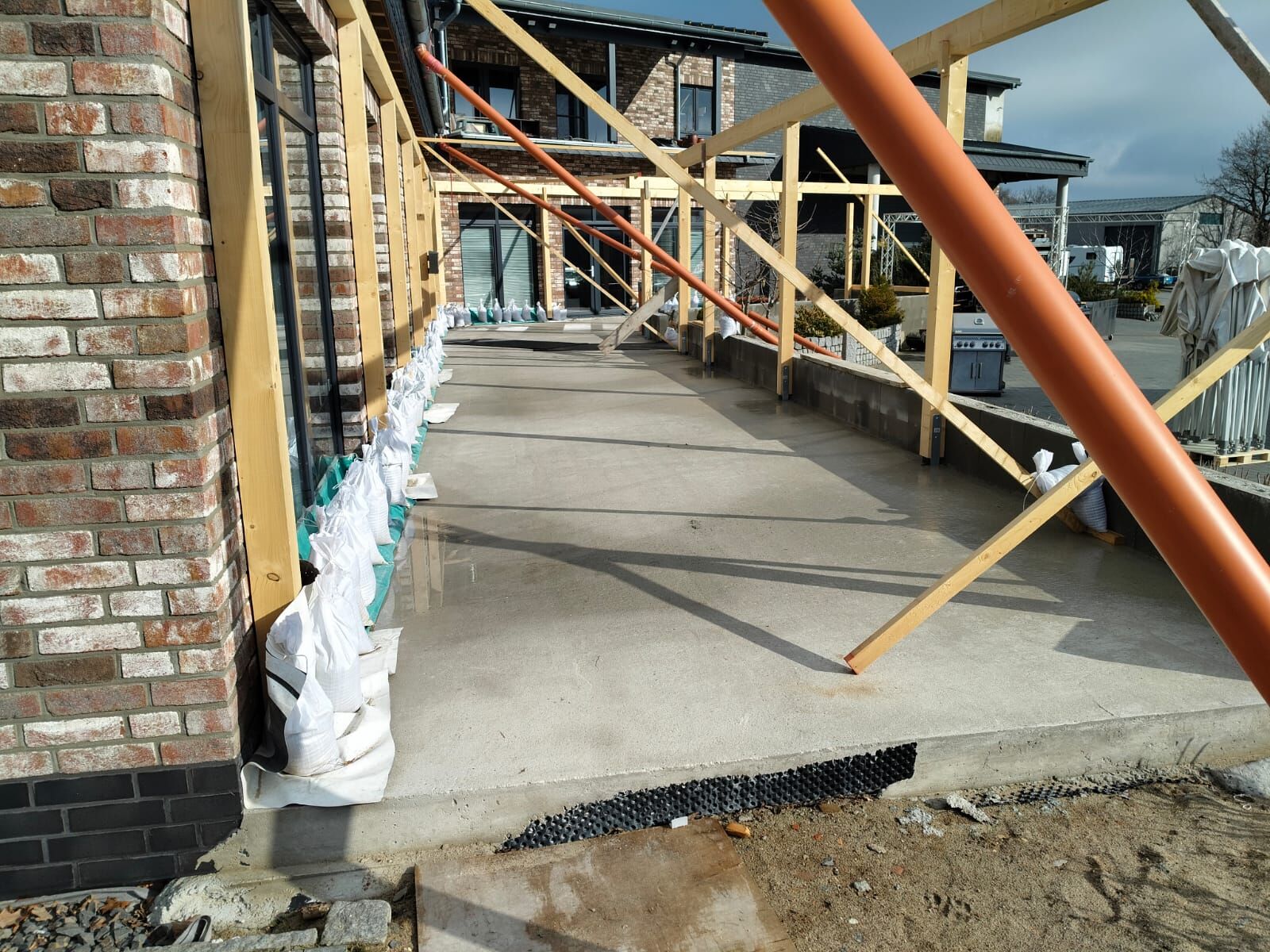 Bauphase einer überdachten Terrasse mit Stützgerüst aus Holz, Sandsäcken zur Abdichtung an den Fensterfronten und vorbereiteter Betonfläche – Neubau in Kombination mit Klinkerfassade.