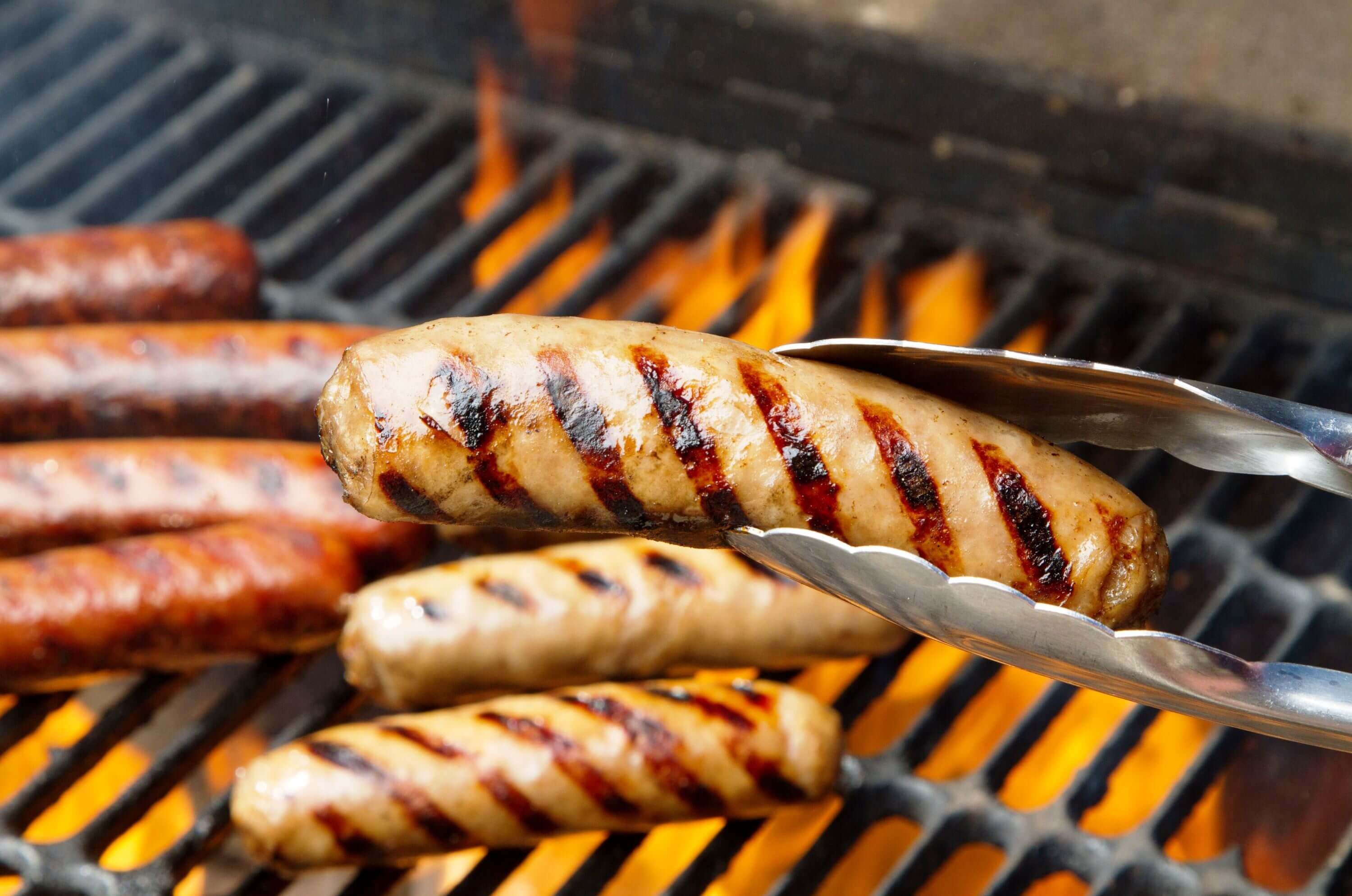 Bratwurst wird auf einem Grill mit Grillzange gewendet, Flammen im Hintergrund.