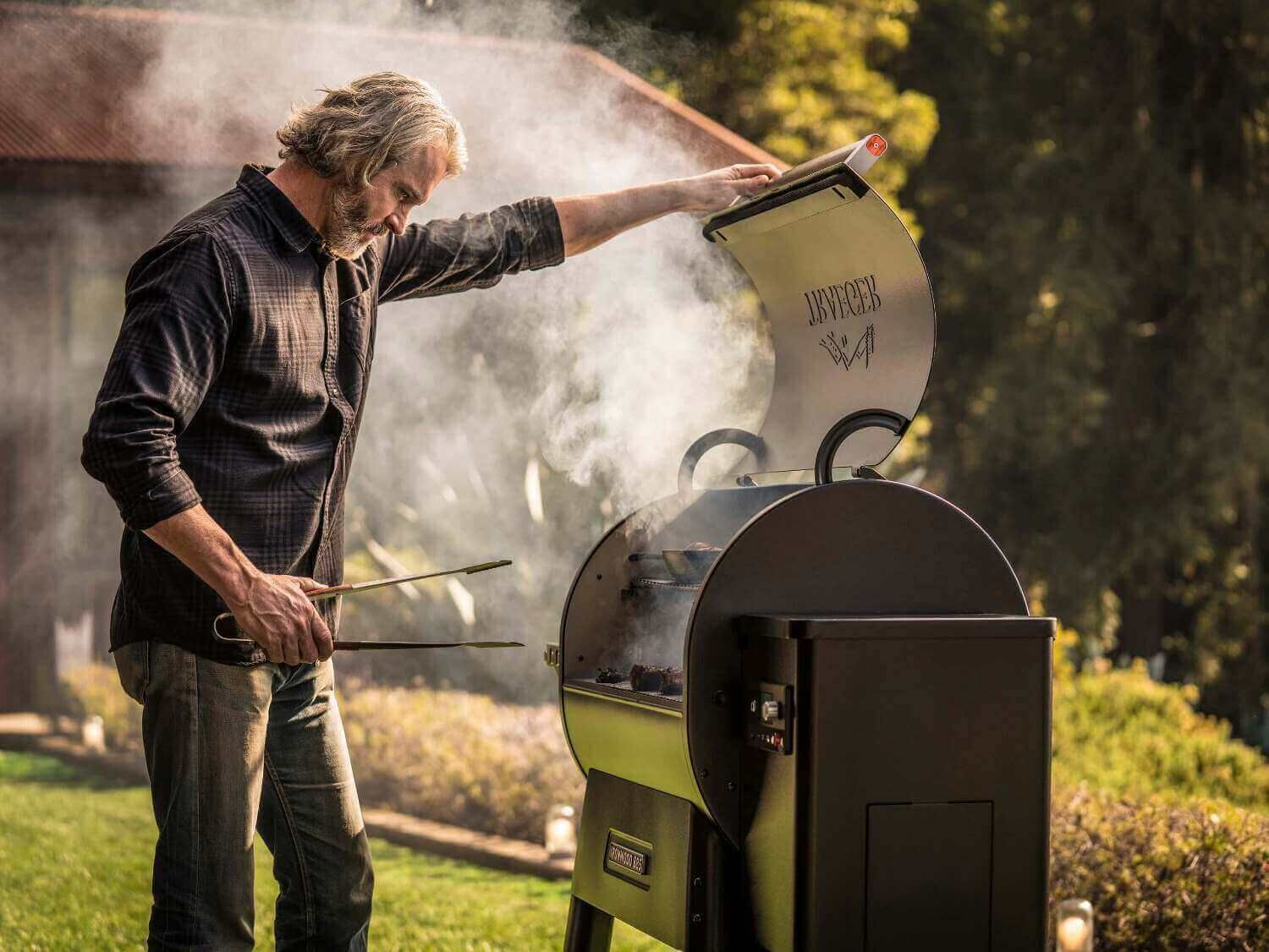 mann-grillt-an-einem-smoker-grill-im-garten-und-öffnet-den-deckel-dichter-rauch-steigt-auf-im-hintergrund-bäume-und-haus-im-abendlicht