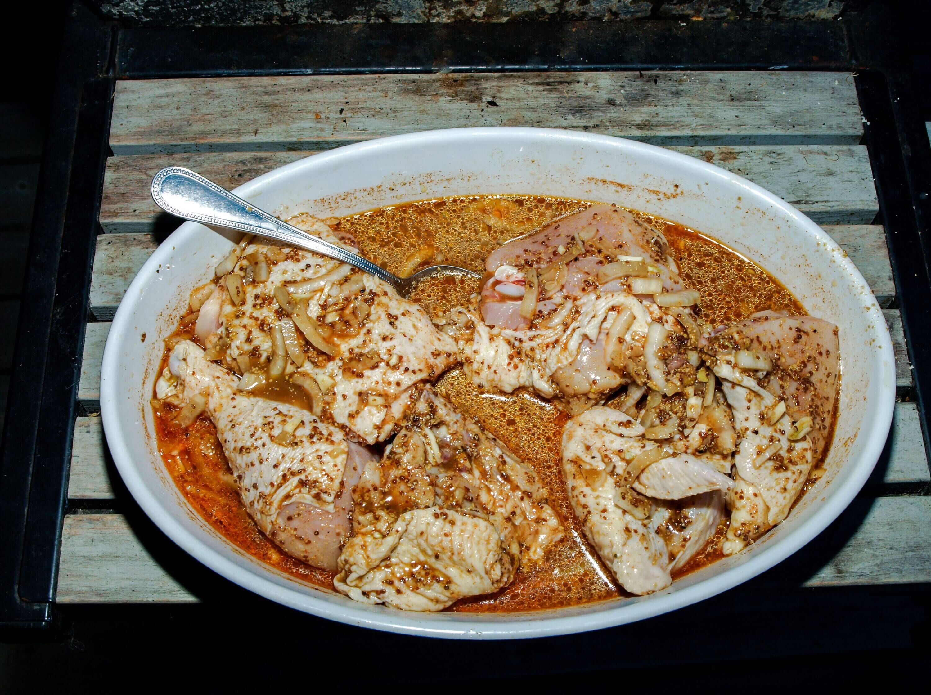 Hähnchenteile in einer würzigen Marinade mit Zwiebeln und Senfkörnern in einer weißen Schale, bereit für den Grill, auf einem rustikalen Holztisch.