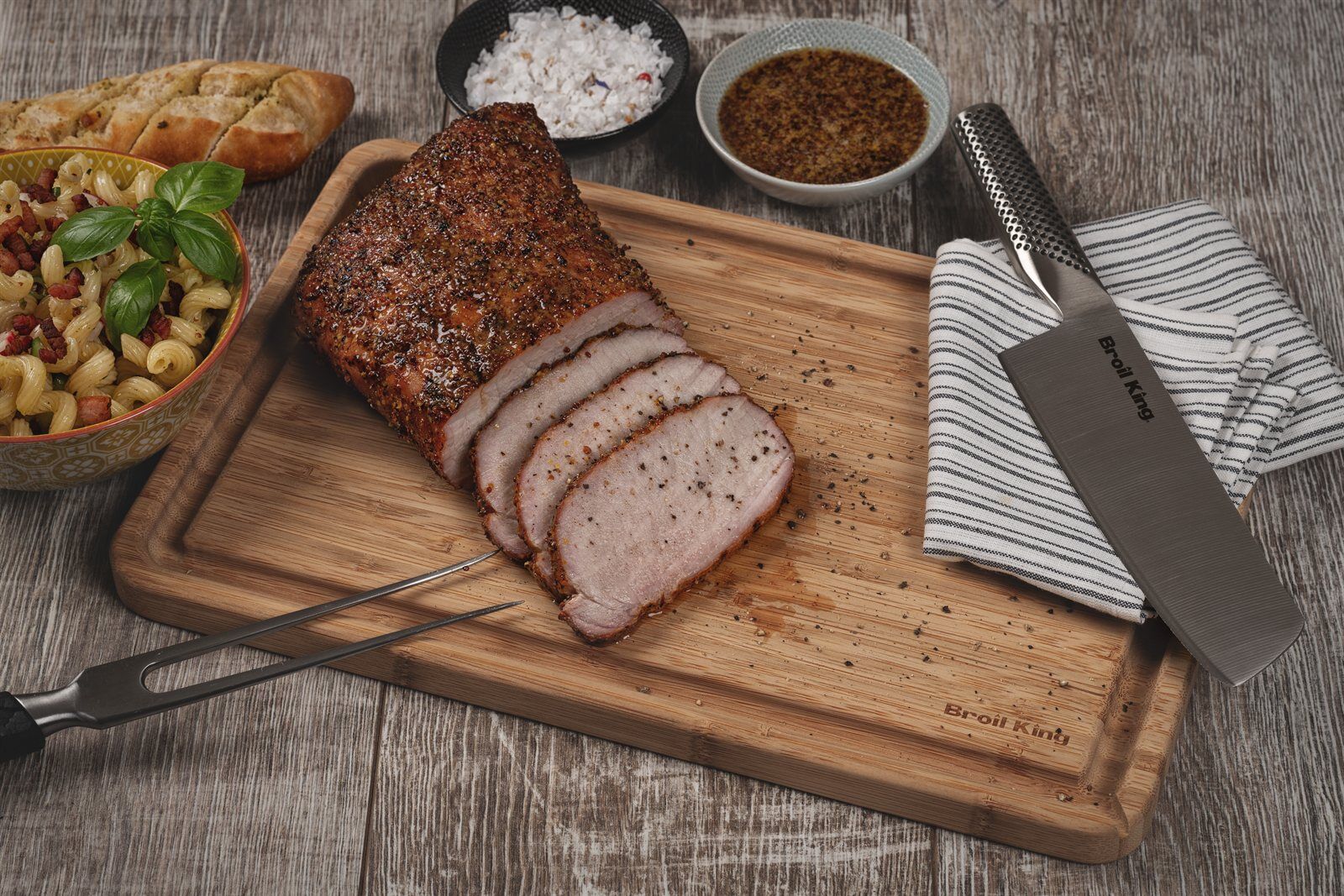 Geräucherter Schweinelachs in Tranchen geschnitten auf dem Holzbrett, daneben Nudelsalat und Baguette