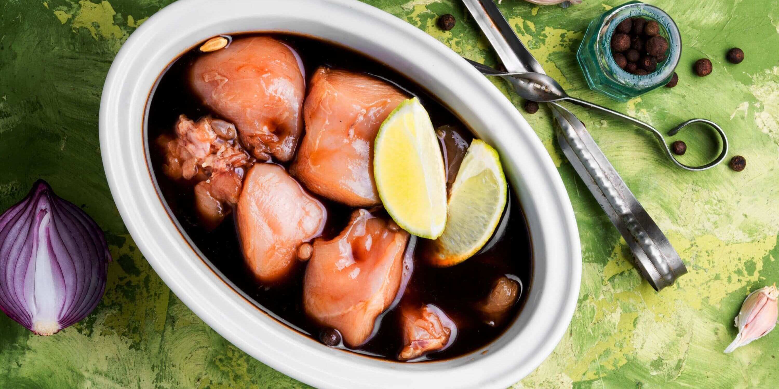 Rohe Hähnchenteile in einer dunklen Marinade mit Limettenstücken, bereit zum Grillen, auf einem grünen Hintergrund mit Zwiebel, Pfefferkörnern und Knoblauch.