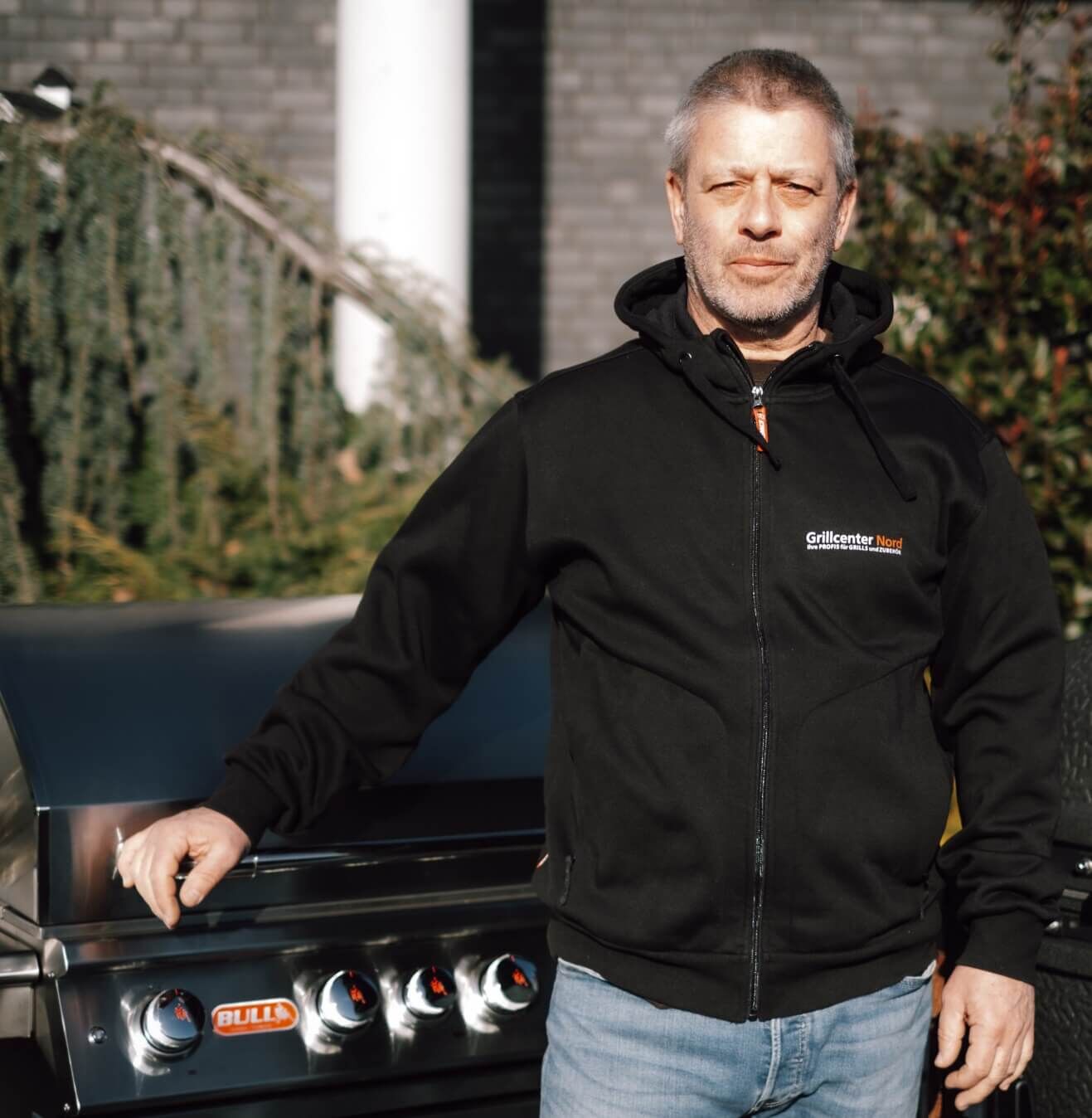 Michael im schwarzen Grillcenter-Nord-Hoodie, eine Hand auf einem Gasgrill