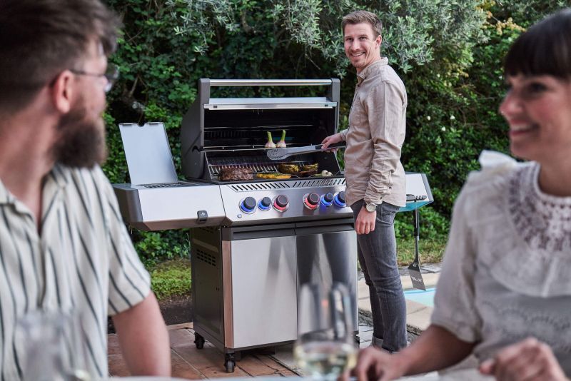 Mann grillt lachend an einem Napoleon Prestige 500 aus Edelstahl, vorne unscharf zwei Gäste am Tisch