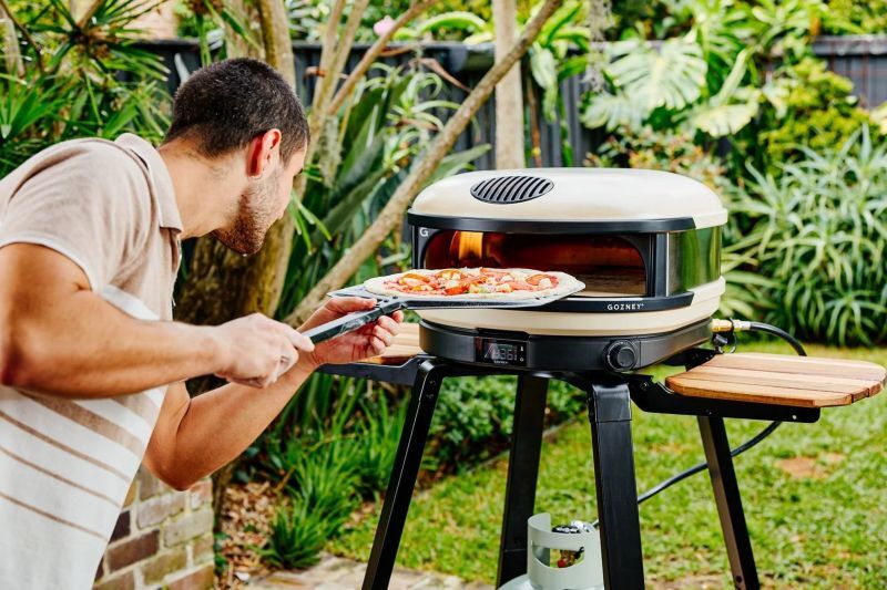 Mann schiebt eine Pizza mit dem Schieber in einen Gozney Pizzaofen auf einem Gestell im Garten