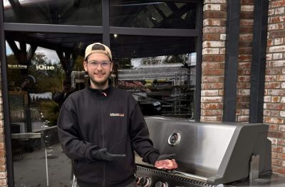 Julius mit Kappe und schwarzen Handschuhen zeigt neben einem Edelstahl-Gasgrill ein rohes Steak