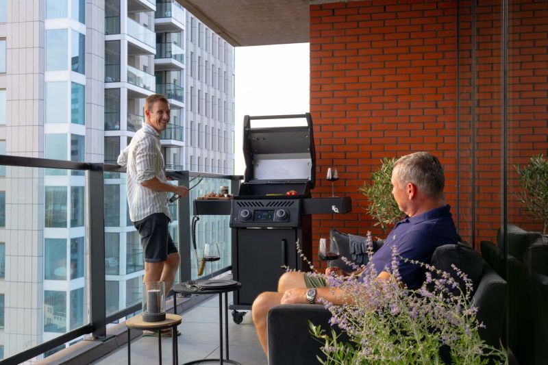 Mann grillt auf dem Hochhausbalkon am Napoleon Elektrogrill, ein Freund sitzt in der Lounge
