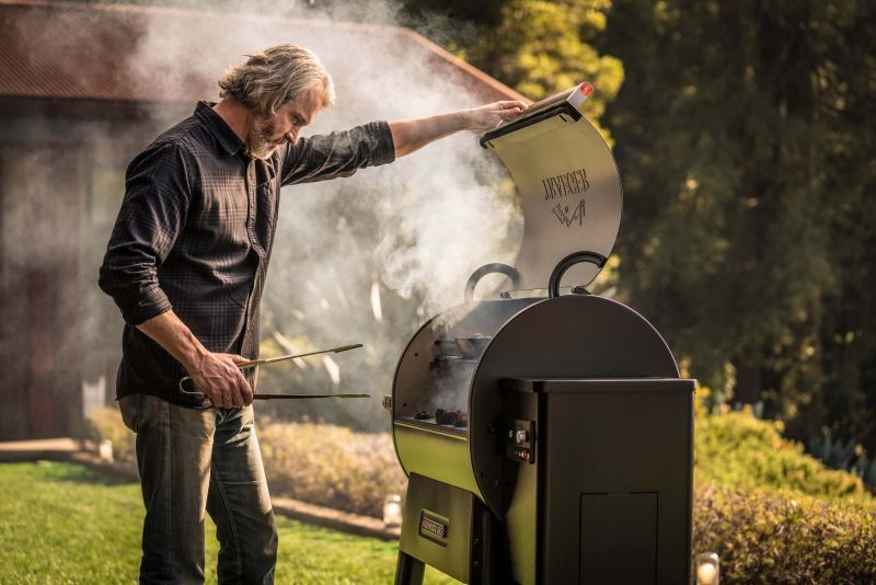 Mann öffnet den Deckel eines Traeger Pelletgrills, Rauch steigt im Abendlicht auf