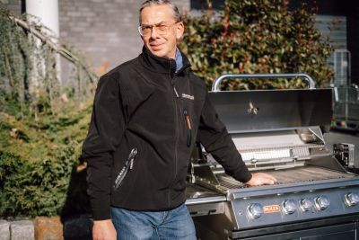 Kay in schwarzer Jacke neben einem Edelstahl-Gasgrill mit geöffnetem Deckel