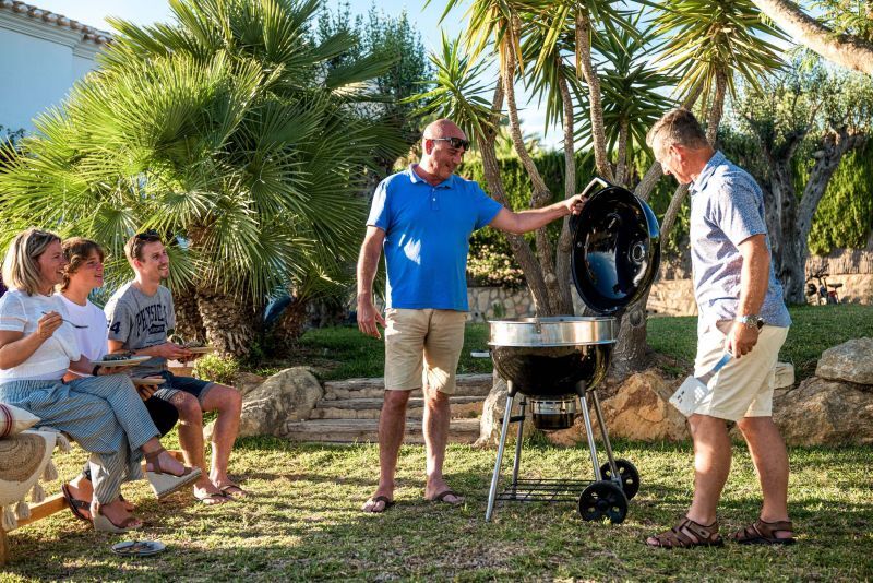 Zwei Männer am Napoleon Kugelgrill mit offenem Deckel im Garten unter Palmen