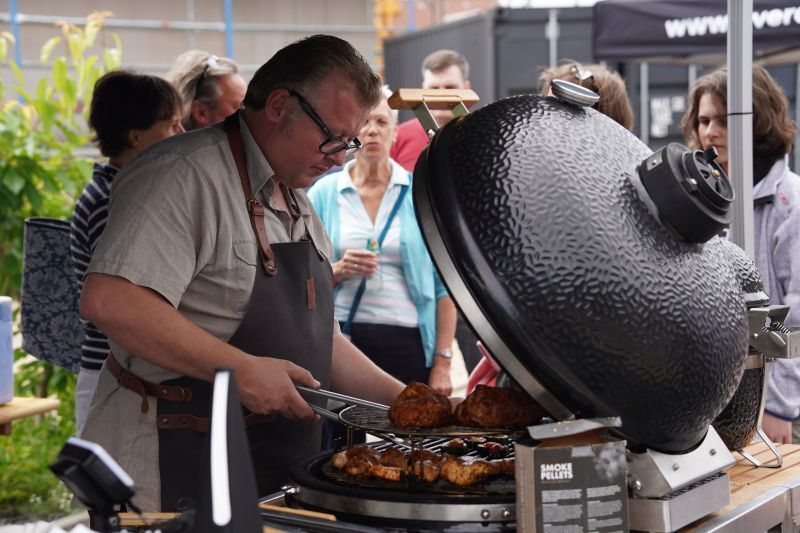 Ein Grillmeister legt Hähnchen auf dem Keramikgrill um, die Gäste stehen dahinter und schauen zu