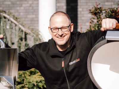 Lars lacht in schwarzer Grillcenter-Nord-Jacke, links und rechts je ein Grill